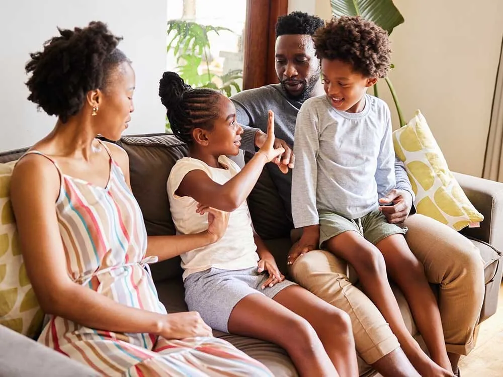 A family of four sit together on a sofa, with a young girl raising her hand toward her father.